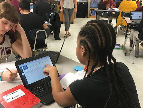 Students in a classroom pointing at a laptop screen.