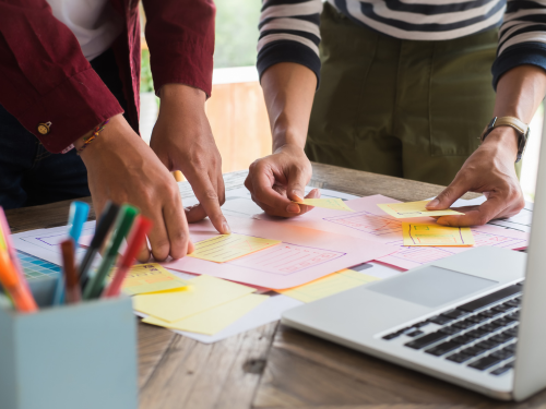 Image: Two pairs of hands pointing at Post-it notes on a table with a laptop computer and a container with pens.