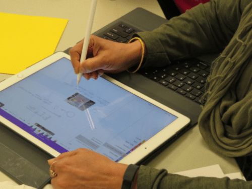 Person using a stylus on a tablet while working at a desk with a laptop and notes nearby.