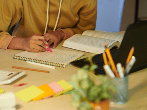 Person studying at a desk, writing notes with a pink highlighter beside an open book, laptop, and colorful sticky notes.